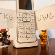 Zwei Lego-Figuren halten Schilder mit "TKG" und "UWG" hoch und stehen neben einem Telefon.