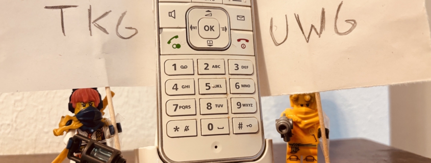 Zwei Lego-Figuren halten Schilder mit "TKG" und "UWG" hoch und stehen neben einem Telefon.